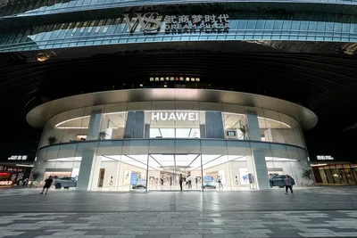 The curved glass frontage of a Huawei store at the WS Dream Plaza shopping mall in Wuhan at night, with cars on display inside and shoppers on the plaza outside