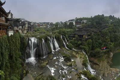 The two-tier waterfall at Furong Town dropping through the gorge, with Tujia stilt houses on the left and the old town built along the cliff above
