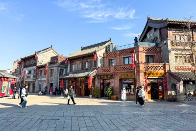 A row of traditional grey-brick shopfronts with red lanterns and carved wooden windows on a paved square at Luoyi Ancient City in Luoyang, with a few visitors in period dress walking past (Photo: Windmemories via Wikimedia Commons)