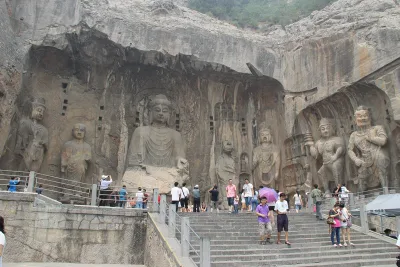 O buda colossal Vairocana e as figuras que o acompanham esculpidos no penhasco do templo Fengxian, nas grutas de Longmen, com visitantes nas escadas abaixo (Photo: Gary Todd via Wikimedia Commons)