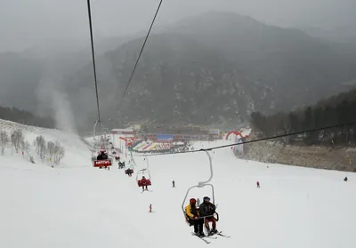 Skiers riding a chairlift above a wide groomed slope at Wanlong ski resort near Chongli, with a crowd gathered at the base station and grey forested ridges beyond (Photo: 黑夜有灯 via Wikimedia Commons)