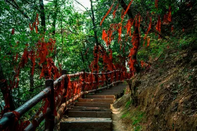 Stone steps climbing through trees hung with red prayer ribbons, beside a wooden handrail on a forest trail in Zhangjiajie